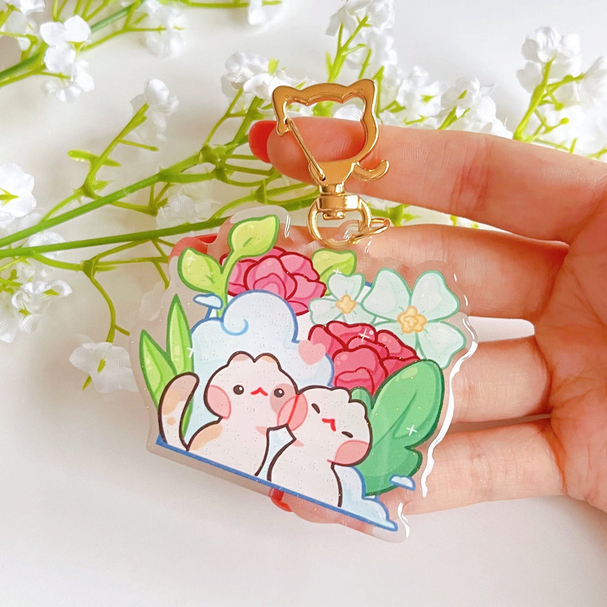A glittery acrylic charm being held in front of some flowers. The design has 2 cats in front of some scenery and the charm has a cat shaped gold keychain accessory.