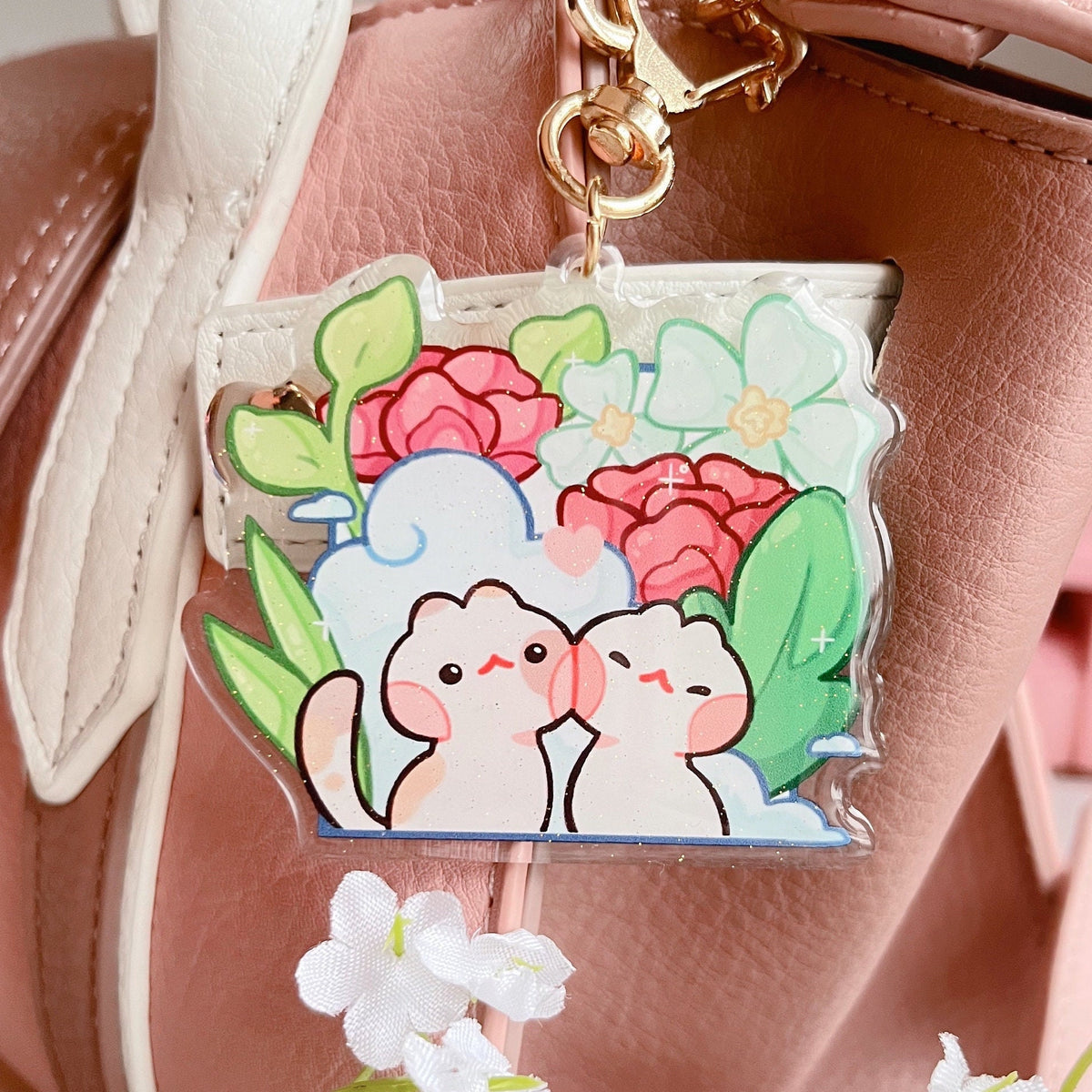 A glittery acrylic charm attached to a bag. The design has 2 cats in front of some scenery and the charm has a cat shaped gold keychain accessory clipped to the bag.