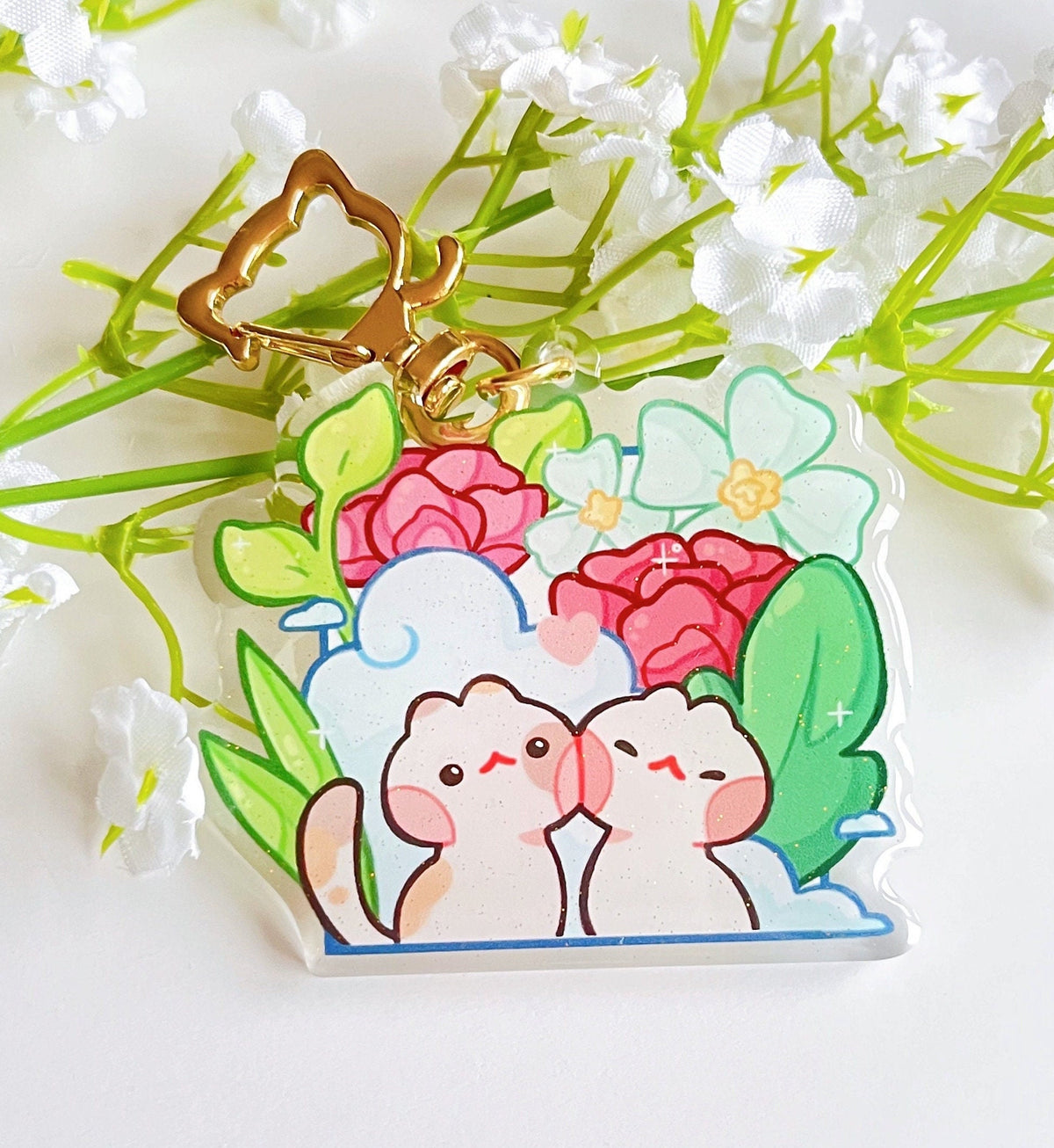 A glittery acrylic charm in front of some flowers. The design has 2 cats in front of some scenery and the charm has a cat shaped gold keychain accessory.