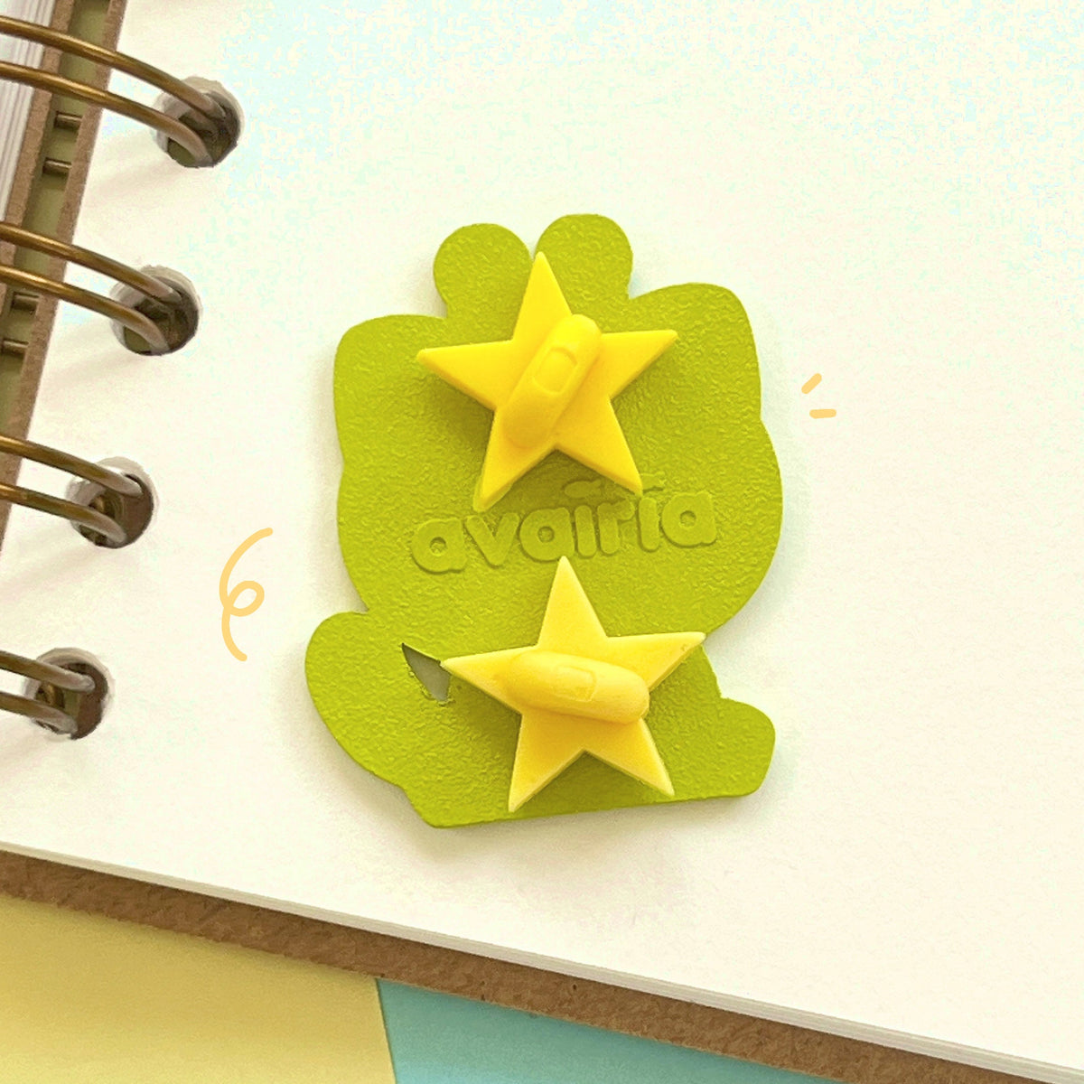 The back of the enamel pin. It has 2 yellow star rubber clasps and a backstamp that reads "avairia".