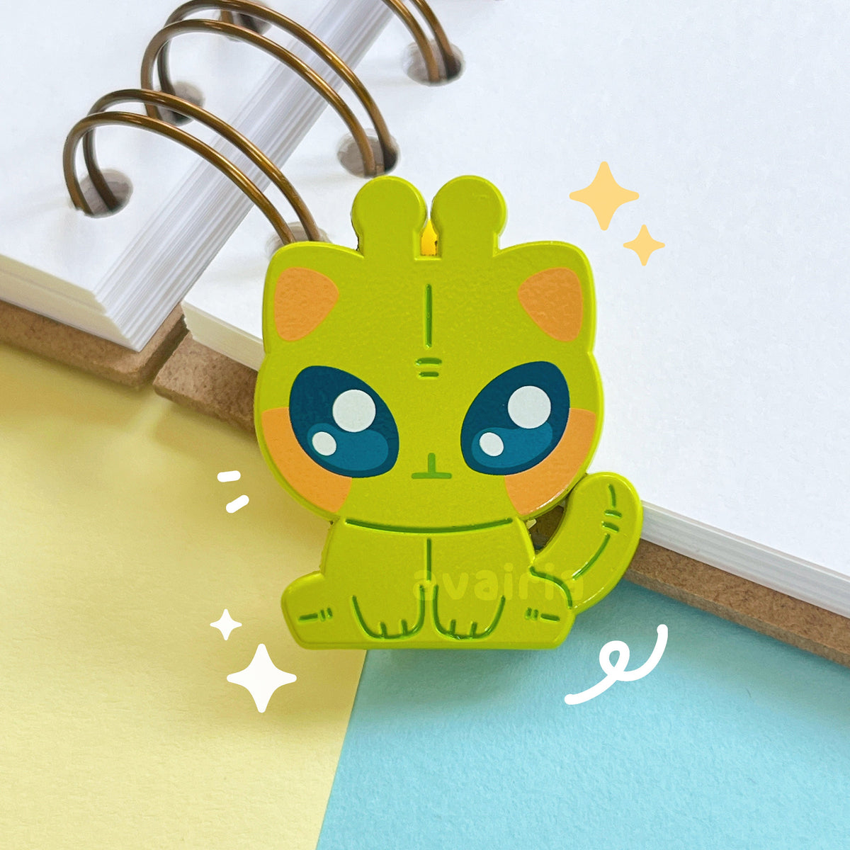 A close up of the enamel pin, it is a lime green alien cat with pink cheeks and ears and blue shiny eyes. The details of the pin are screen printed and some parts are a dark green enamel.