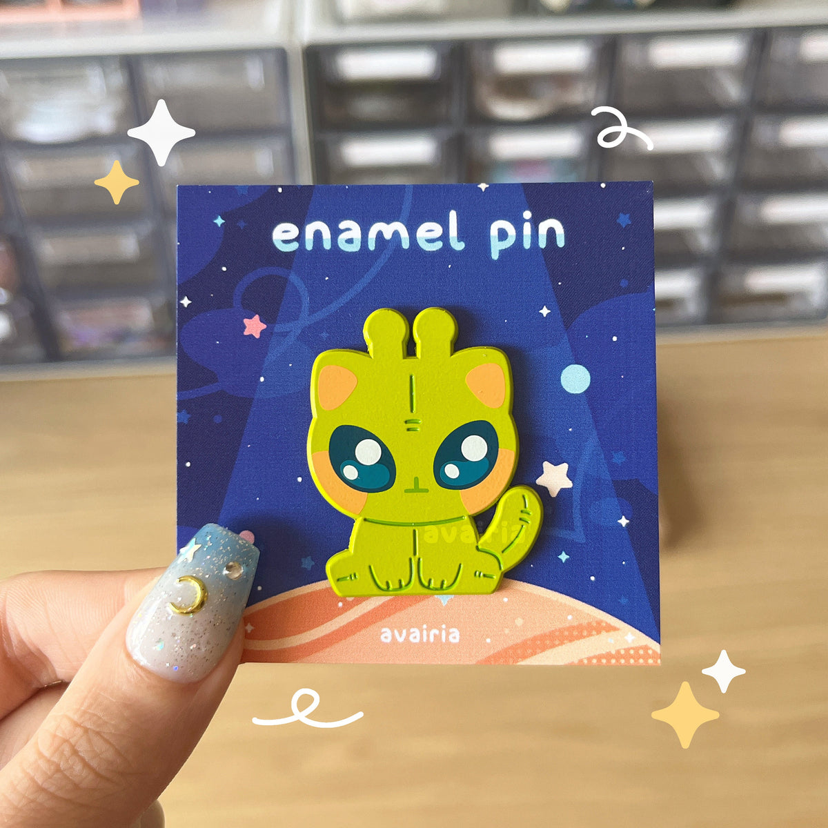 A green alien cat sitting down enamel pin on a dark blue backing card with stars and a planet.