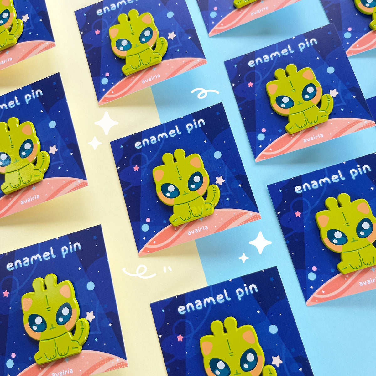 Multiple green alien cat enamel pins attached to their backing cards.