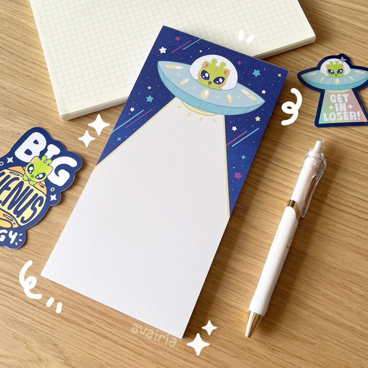 A large sized notepad, the design has a alien cat in a UFO on a darker blue background with colourful stars. The UFO is shooting down a light beam.