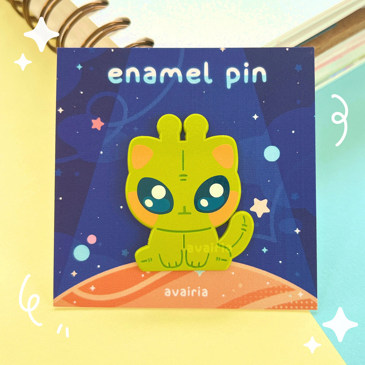 A green alien cat sitting down enamel pin on a dark blue backing card with stars and a planet.