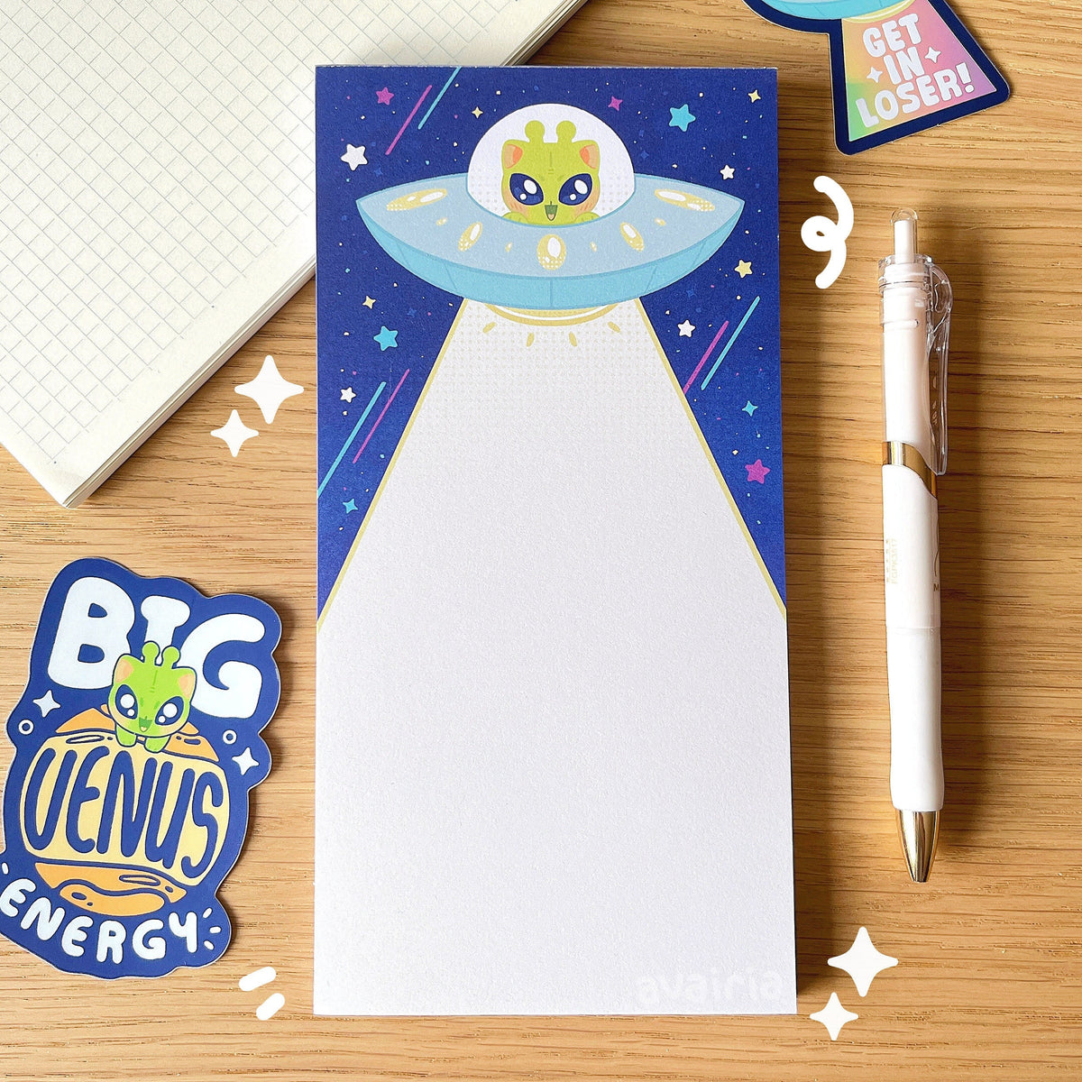 A Large sized notepad, the design has a alien cat in a UFO on a darker blue background with colourful stars. The UFO is shooting down a light beam.