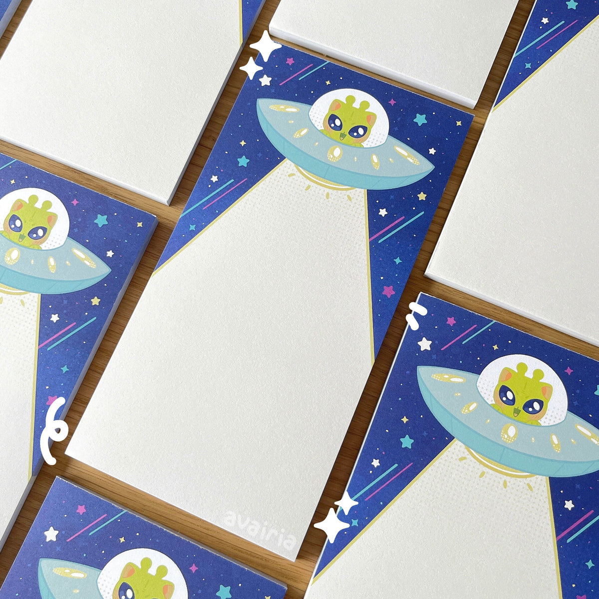 Multiple notepads, the design has a alien cat in a UFO on a darker blue background with colourful stars. The UFO is shooting down a light beam.