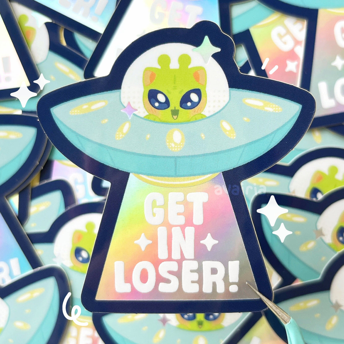 A vinyl sticker of an alien cat inside a UFO. A beam is shooting down from the UFO and inside the beam there is white text that reads "GET IN LOSER!". The beam and stars on the sticker have a holographic effect.