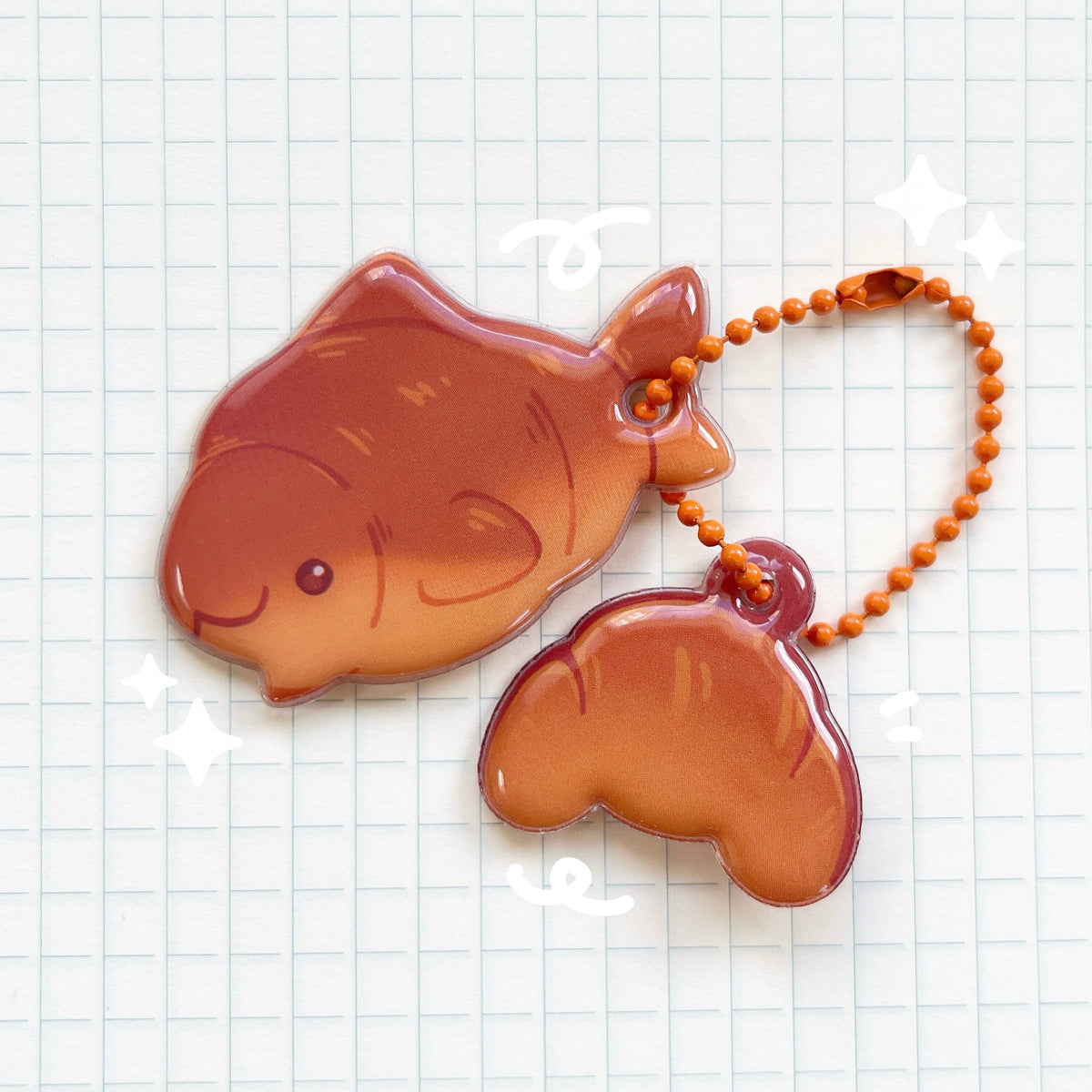 A PVC puffy charm with two charms, the top left is a croissant shark, the bottom right is a croissant. They are attached to an orange ball chain accessory.