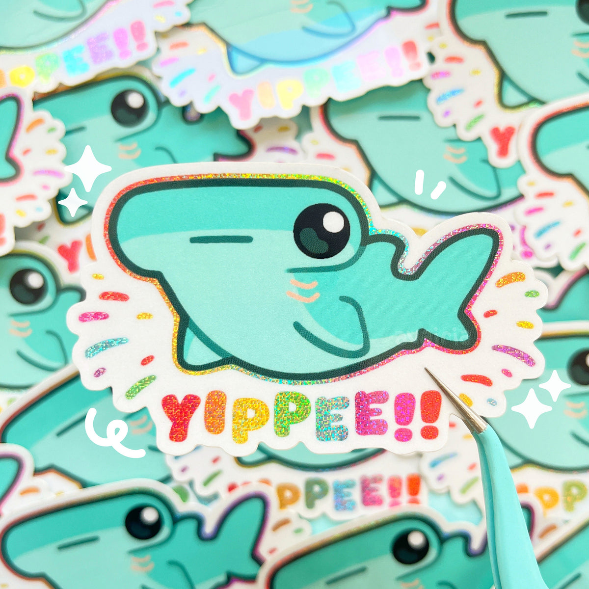 A sticker of a turquoise hammerhead shark with rainbow holographic pixie dust effects surrounding it, the text below reads "YIPPEE!!" in rainbow colours.