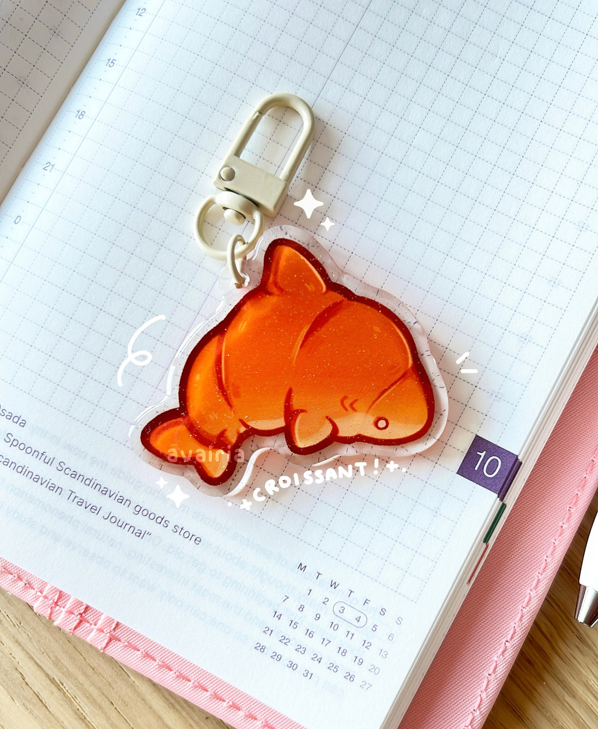 An acrylic charm of a croissant shark with a gold glitter effect. The charm has a beige clasp accessory.
