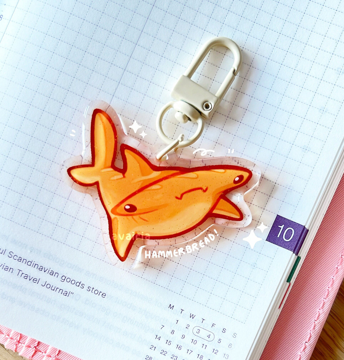 An acrylic charm of a Hammerbread shark with a gold glitter effect. The charm has a beige clasp accessory.
