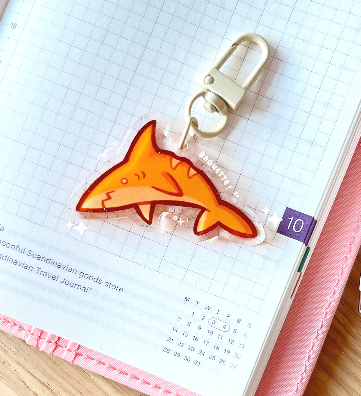 An acrylic charm of a baguette shark with a gold glitter effect. The charm has a beige clasp accessory.