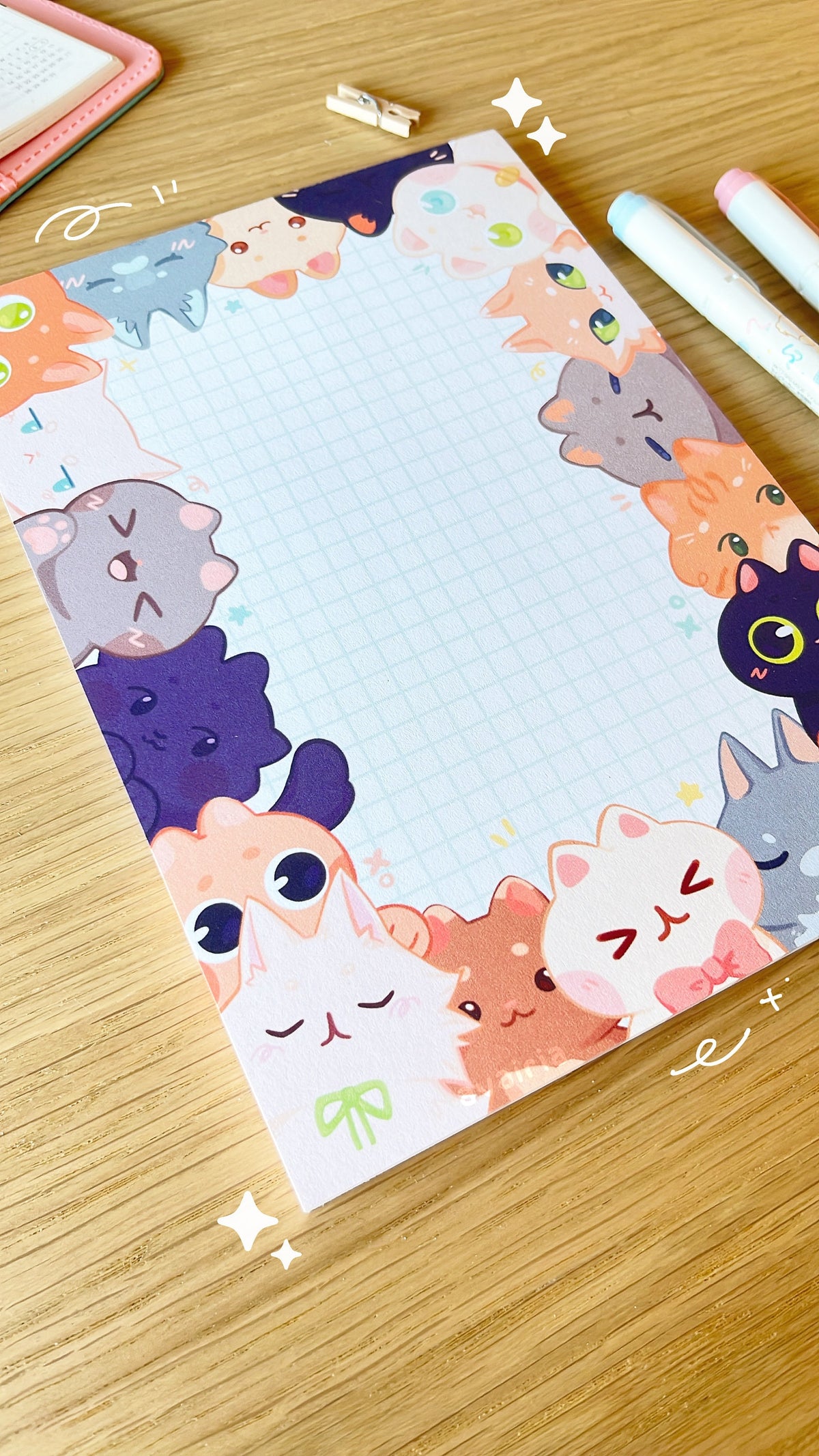 A large notepad, the design has a border of different types of cats peaking out from the sides, the centre is a white background with a blue grid design.