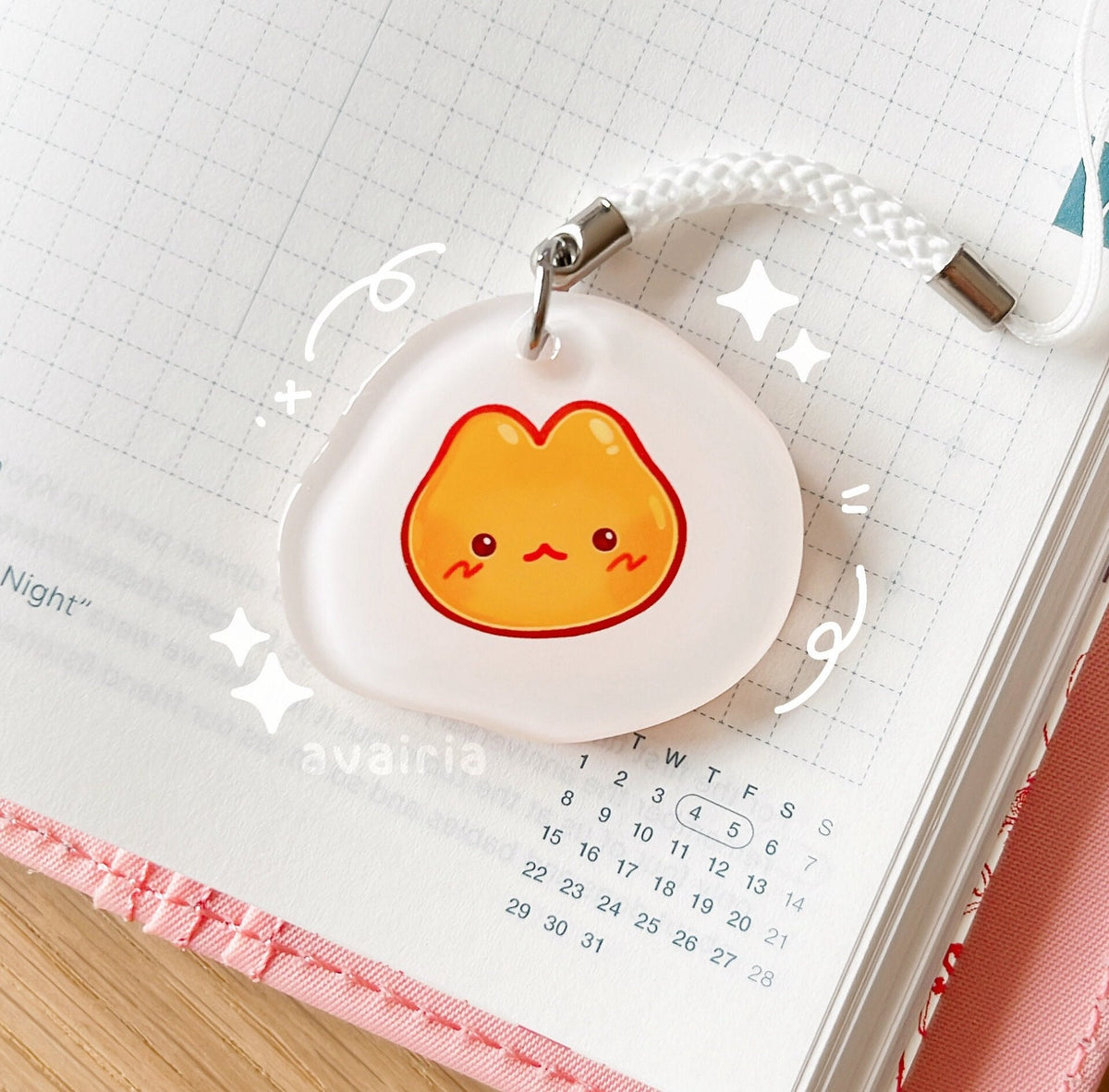 A small acrylic charm of an egg themed cat. It has a white phone strap accessory.
