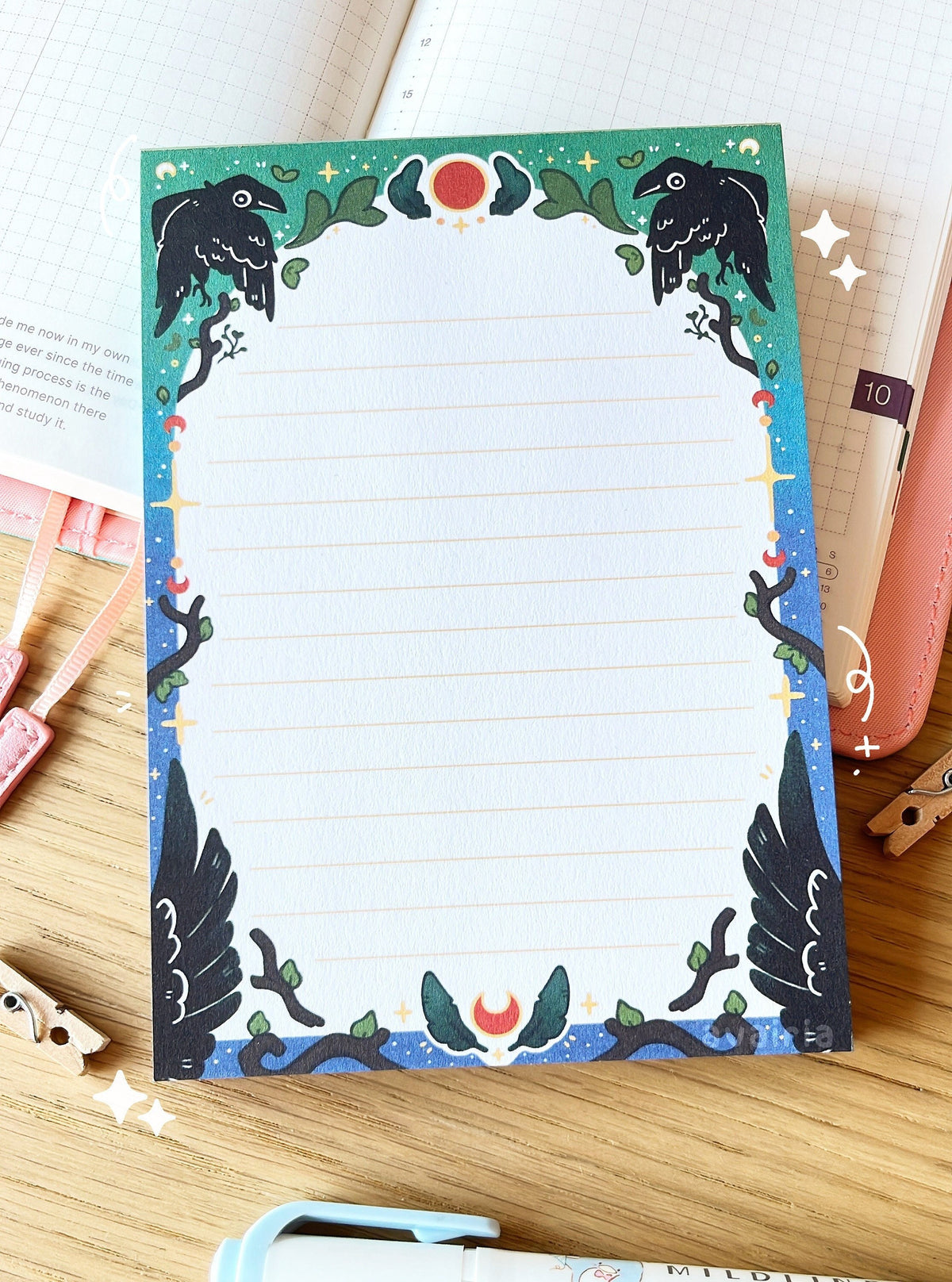 The notepad has an arched green to blue border with a crow, feather and twig design with small star details. The inside of the border is white with small yellow lines.