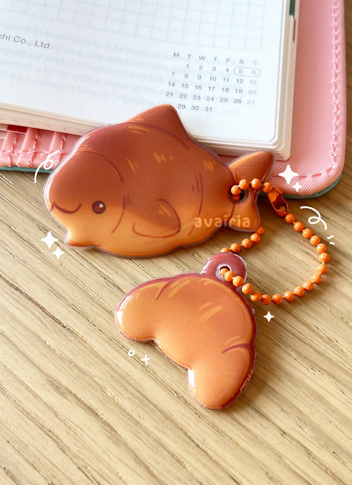 A PVC puffy charm with two charms, the top is a croissant shark, the bottom is a croissant. They are attached to an orange ball chain accessory.