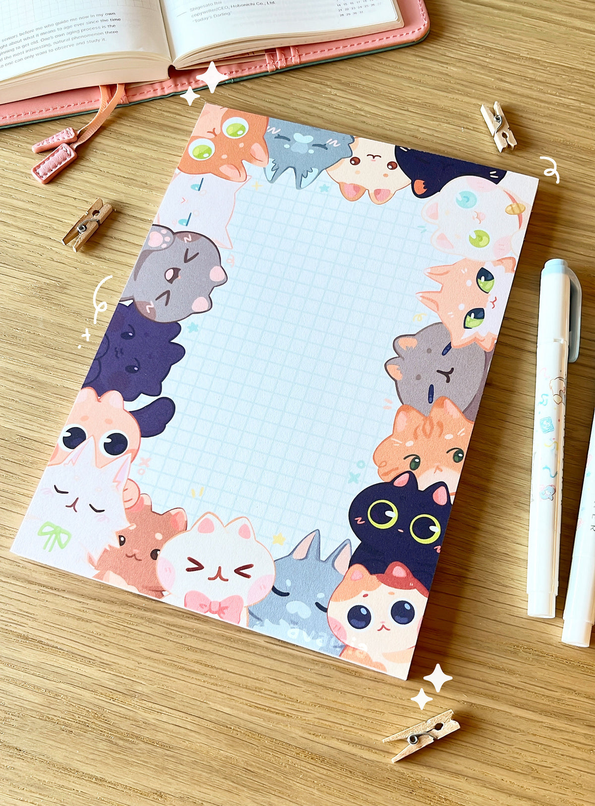 A large notepad, the design has a border of different types of cats peaking out from the sides, the centre is a white background with a blue grid design.