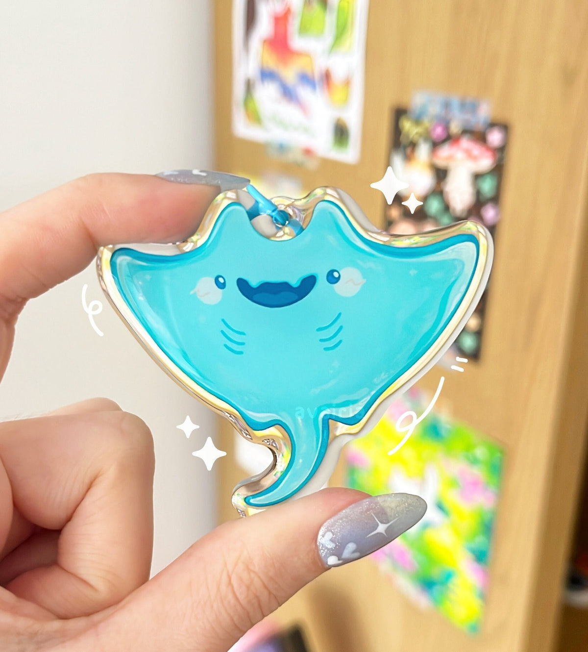 A turquoise manta ray acrylic charm being held up. It has a blue keychain clip accessory.